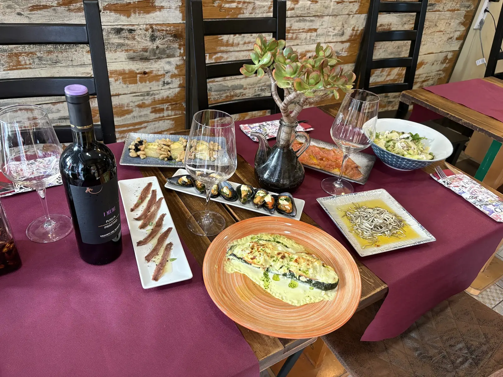 Mesa puesta con mantel burdeos: botella de vino tinto Primitivo de Puglia, anchoas, mejillones gratinados, ensalada y calabacín gratinado